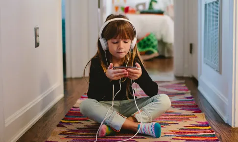 Child sitting cross-legged on the floor — using her phone and headphones